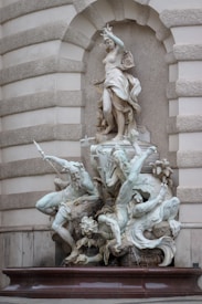 A marble sculpture depicting a mythological scene with a prominently positioned female figure standing over several other figures and sea creatures. The figures are detailed with flowing garments and dynamic poses, seemingly engaged in a dramatic interaction. The background is an ornate architectural niche with textured stonework.