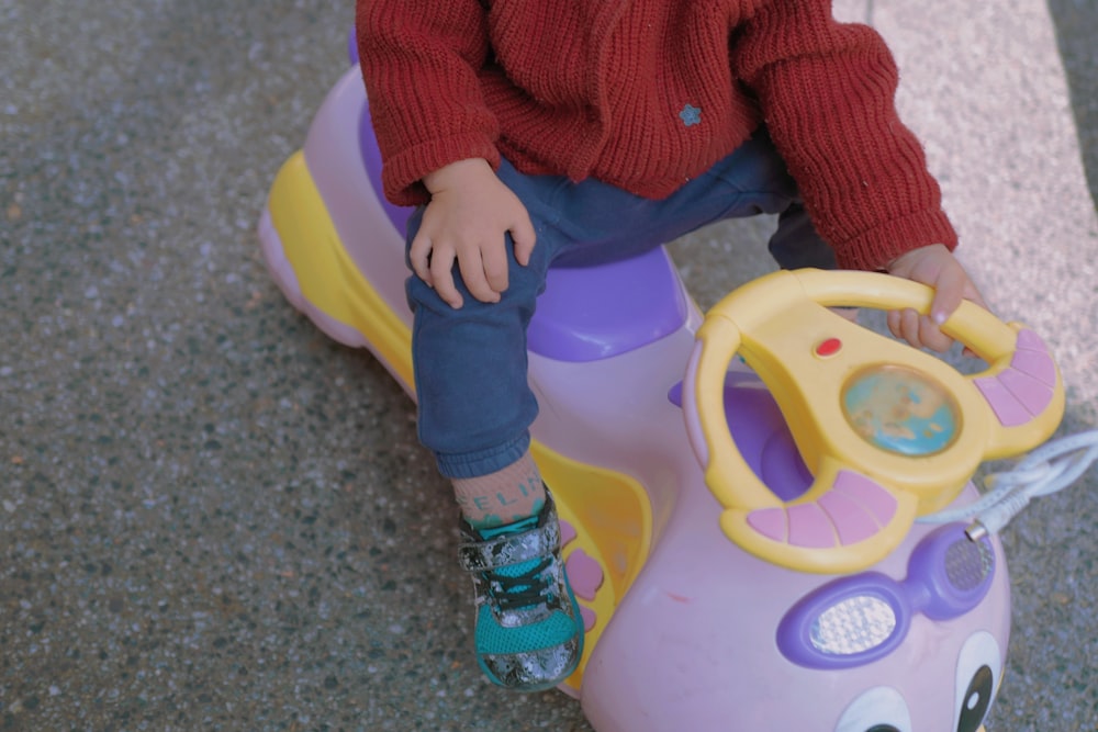 child in red sweater and blue pants sitting on yellow and green plastic toy