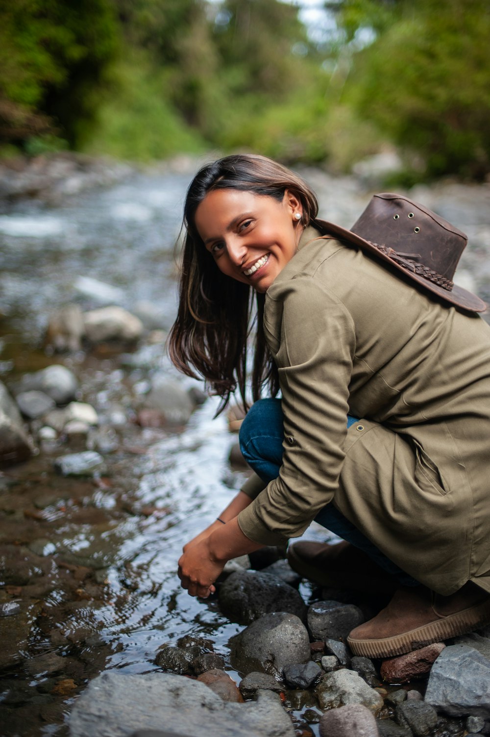 woman in green jacket and blue denim jeans sitting on rock near river during daytime