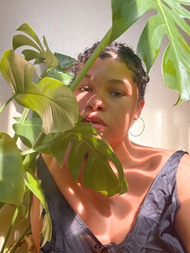 A person partially obscured by large green leaves, standing in a softly lit environment. Their expression is calm and introspective, with sunlight casting gentle shadows across their face and clothing.