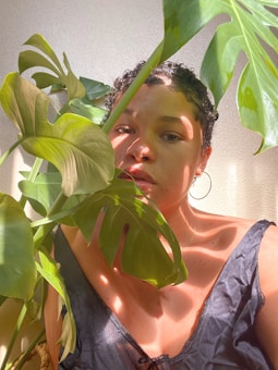 A person partially obscured by large green leaves, standing in a softly lit environment. Their expression is calm and introspective, with sunlight casting gentle shadows across their face and clothing.