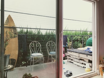 A cozy outdoor balcony features a sliding glass door leading to a space decorated with rustic and casual elements. A surfboard is propped against the railing, next to two metal chairs with decorative backrests. The seating area includes a makeshift couch made from wooden pallets, topped with cushions. Various plants add a touch of greenery, and there is a pair of sandals on the floor. The scene is framed by tall green shrubs and a cloudy sky.