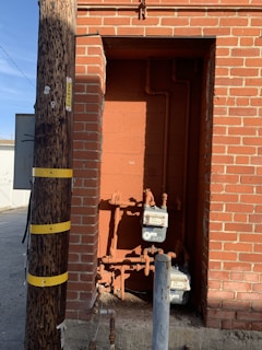 A brick wall with a recessed alcove containing utility meters and pipes. Adjacent to the wall is a wooden utility pole with yellow bands and tags. The ground is concrete, and a metal post is in front of the alcove.