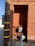 A brick wall with a recessed alcove containing utility meters and pipes. Adjacent to the wall is a wooden utility pole with yellow bands and tags. The ground is concrete, and a metal post is in front of the alcove.