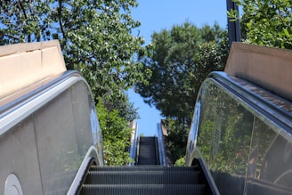 An outdoor escalator surrounded by lush green trees and plants, leading upwards into a clear blue sky. The perspective is from the bottom looking up, creating a sense of ascent and openness.