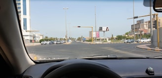 View from inside a car looking out at an intersection with multiple lanes. Traffic lights are present, and several cars are visible, waiting for the signal to change. Surrounding the intersection are buildings, including a tall white building on the left and other shorter buildings on the right. There are also billboards on the roadside.