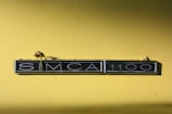 A yellow background featuring a classic car badge with the text 'SIMCA 1100'. The badge has some small floral elements resting on top of it, suggesting a nostalgic or vintage theme.
