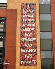 A mural with colorful abstract patterns features text calling for world peace, universal healthcare, and the end of oppression, prejudice, and poverty. The mural is located on the side of a brick building, with a no entry traffic sign visible nearby.