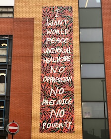 A mural with colorful abstract patterns features text calling for world peace, universal healthcare, and the end of oppression, prejudice, and poverty. The mural is located on the side of a brick building, with a no entry traffic sign visible nearby.