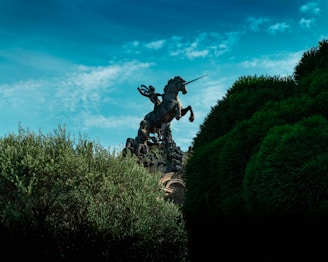 A majestic statue of a warrior riding a rearing unicorn is partially obscured by lush green foliage. The sky above is a vibrant blue with wispy clouds, contributing to a dramatic and mythical atmosphere.