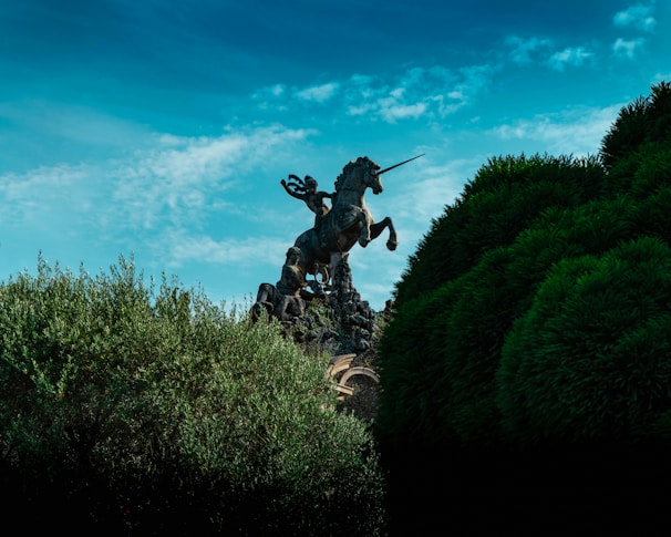 A majestic statue of a warrior riding a rearing unicorn is partially obscured by lush green foliage. The sky above is a vibrant blue with wispy clouds, contributing to a dramatic and mythical atmosphere.
