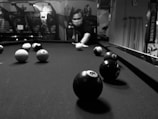 A person wearing a mask is about to strike a cue ball on a pool table. The table is covered in black felt, with several billiard balls distributed across it. The setting appears to be indoors, with various arcade games or machines visible in the background.