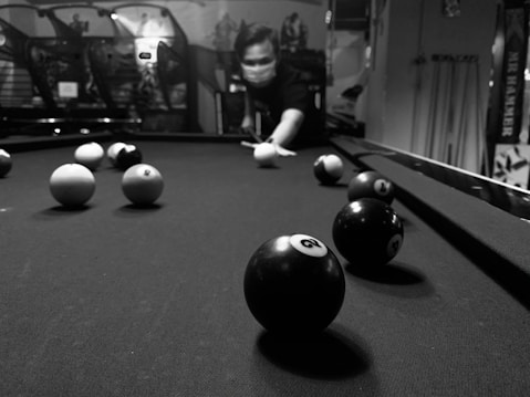 A person wearing a mask is about to strike a cue ball on a pool table. The table is covered in black felt, with several billiard balls distributed across it. The setting appears to be indoors, with various arcade games or machines visible in the background.