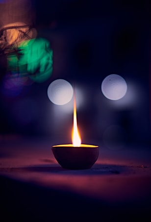 A single lit oil lamp, placed on a flat surface, with a steady flame that casts a warm glow. The background is dark, featuring blurred circular lights and abstract shapes, giving a peaceful and contemplative atmosphere.