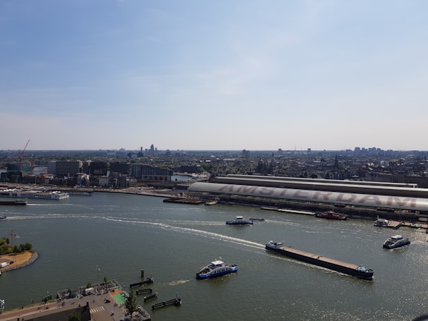 A panoramic view of a city waterfront with multiple boats on a wide river. On the opposite side, a large building with the word 'AMSTERDAM' clearly visible on its roof. The skyline is filled with a mix of modern and older architectural structures under a clear blue sky.