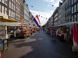 A bustling street market lined with various stalls offering goods such as clothing, food, and other items. The market is situated between tall brick buildings with a clear sky above. A large Dutch flag and colorful pennant flags are strung above the street, adding a festive atmosphere. Several people are walking along the market, browsing through the items.