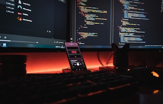 A dark, ambient workspace featuring two computer monitors displaying lines of colorful code. A smartphone with a music player is positioned vertically on the desk, with a glowing orange-red light providing an atmospheric background. Various peripherals like a keyboard and a mouse are partially visible, with a camera lens or similar object on the right.