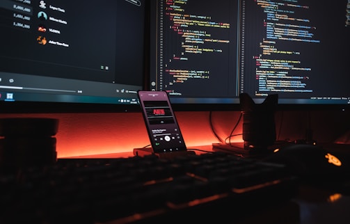 A dark, ambient workspace featuring two computer monitors displaying lines of colorful code. A smartphone with a music player is positioned vertically on the desk, with a glowing orange-red light providing an atmospheric background. Various peripherals like a keyboard and a mouse are partially visible, with a camera lens or similar object on the right.