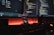 A dark, ambient workspace featuring two computer monitors displaying lines of colorful code. A smartphone with a music player is positioned vertically on the desk, with a glowing orange-red light providing an atmospheric background. Various peripherals like a keyboard and a mouse are partially visible, with a camera lens or similar object on the right.