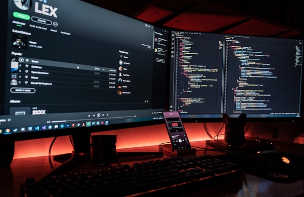 A dual-monitor computer setup displays code on the right screen and a music streaming service on the left. A smartphone on a stand shows a music app, and a mechanical keyboard is visible in front. The setup is illuminated by ambient red lighting.