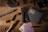 A wooden surface contains various items, including a folding knife, a fabric wallet, playing cards, a cigar, bullet casings, a pen, a ring, a drink, coins, and small cubes resembling dice. The arrangement suggests a rustic or adventurous theme.