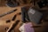 A wooden surface contains various items, including a folding knife, a fabric wallet, playing cards, a cigar, bullet casings, a pen, a ring, a drink, coins, and small cubes resembling dice. The arrangement suggests a rustic or adventurous theme.