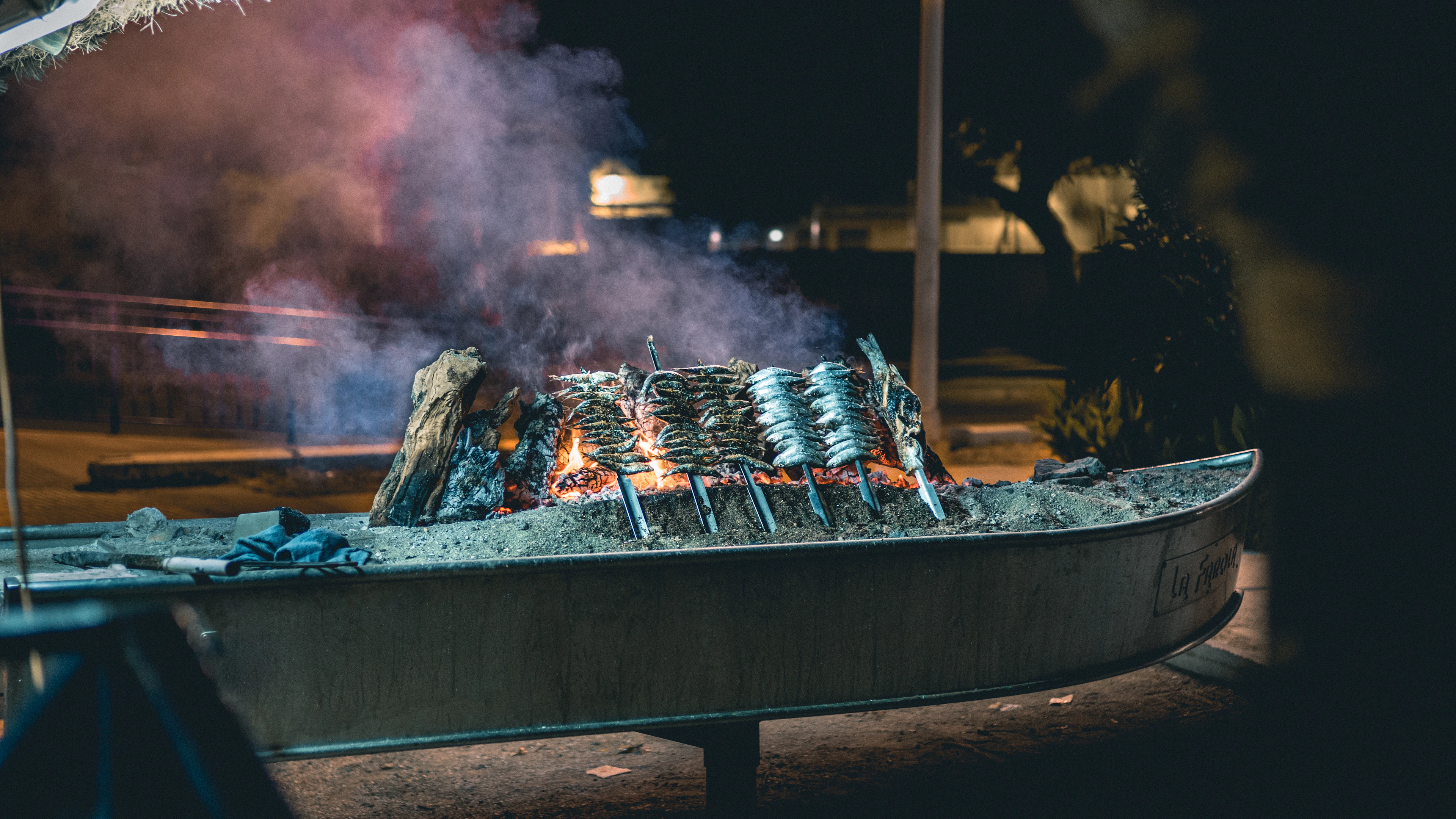 Sardine skewers (espetos) grilling over open flames in a boat-shaped grill at night.