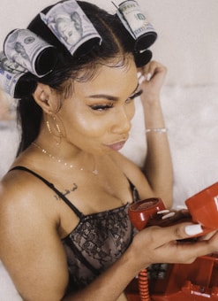 A woman with dollar bills styled in her hair like curlers is holding a red vintage telephone receiver. She is wearing a black lace outfit and hoop earrings, with notable makeup and accessories.