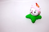 A plush toy with a cute face and bright colors, featuring a white head with large eyes and a smiling expression. It wears a pink hat with what appears to be a red point or flame. The body is shaped like a green starfish or legs.