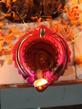 A decorative, intricately designed candle holder with a vibrant red and pink hue, surrounded by scattered orange marigold petals. The candle is lit, emitting a warm glow that enhances the colors surrounding it.