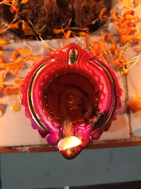 A decorative, intricately designed candle holder with a vibrant red and pink hue, surrounded by scattered orange marigold petals. The candle is lit, emitting a warm glow that enhances the colors surrounding it.