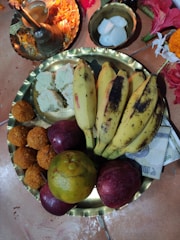A brass plate contains a variety of fruits including bananas, apples, and an orange. Sweets and what appears to be coconut pieces are also present. Nearby, a small metal dish with white circular items is visible, along with several vibrant flowers. There's a small brass lamp with marigold petals and incense sticks.