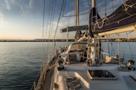 A sailing yacht with a wooden deck, ropes, and masts is anchored on calm water. The horizon in the distance shows a coastline with buildings and a slightly cloudy sky at sunset.