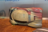 A large, sculpted object sits on a wooden floor. It appears to be an aerodynamic car shell prototype or model with multiple colors, including shades of white, beige, and red. There are some hand-drawn markings and measurements visible on its surface. The background is a plain wall with blue text partially visible.