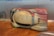 A large, sculpted object sits on a wooden floor. It appears to be an aerodynamic car shell prototype or model with multiple colors, including shades of white, beige, and red. There are some hand-drawn markings and measurements visible on its surface. The background is a plain wall with blue text partially visible.