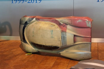 A large, sculpted object sits on a wooden floor. It appears to be an aerodynamic car shell prototype or model with multiple colors, including shades of white, beige, and red. There are some hand-drawn markings and measurements visible on its surface. The background is a plain wall with blue text partially visible.