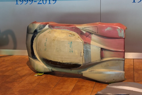 A large, sculpted object sits on a wooden floor. It appears to be an aerodynamic car shell prototype or model with multiple colors, including shades of white, beige, and red. There are some hand-drawn markings and measurements visible on its surface. The background is a plain wall with blue text partially visible.
