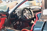 A luxury sports car interior featuring a sleek, high-tech dashboard with a carbon fiber finish. The steering wheel is prominently displayed, surrounded by a variety of meticulously designed control panels. Red accents highlight the seats and parts of the door, contributing to the contemporary design aesthetic.