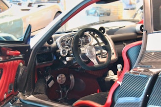 A luxury sports car interior featuring a sleek, high-tech dashboard with a carbon fiber finish. The steering wheel is prominently displayed, surrounded by a variety of meticulously designed control panels. Red accents highlight the seats and parts of the door, contributing to the contemporary design aesthetic.