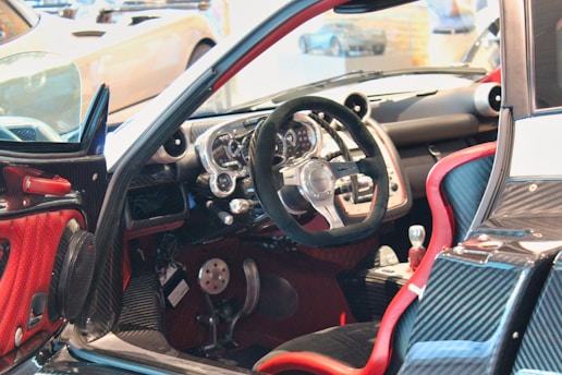 A luxury sports car interior featuring a sleek, high-tech dashboard with a carbon fiber finish. The steering wheel is prominently displayed, surrounded by a variety of meticulously designed control panels. Red accents highlight the seats and parts of the door, contributing to the contemporary design aesthetic.