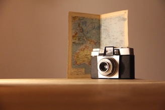 A vintage camera is placed on a wooden surface with an open map standing behind it. The map appears to show geographical details, possibly of Australia and surrounding areas. The scene is softly lit, casting gentle shadows.
