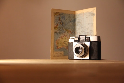 A vintage camera is placed on a wooden surface with an open map standing behind it. The map appears to show geographical details, possibly of Australia and surrounding areas. The scene is softly lit, casting gentle shadows.