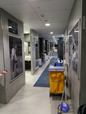 A hallway with gray tiles and blue carpet features cleaning equipment, including a yellow cleaning cart and a mop. There are black and white sports photographs on the walls and a door leading to a restroom area. Overhead, there are ceiling lights providing illumination.