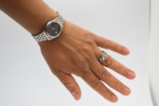 A hand wearing a silver wristwatch with a black watch face and a silver bracelet. Additionally, there are two rings on the ring finger, one with a prominent gemstone and the other a simple band.