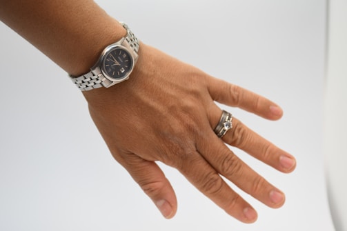 A hand wearing a silver wristwatch with a black watch face and a silver bracelet. Additionally, there are two rings on the ring finger, one with a prominent gemstone and the other a simple band.