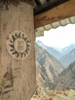 A sun-themed sticker with 'Trippy Hippie' is affixed to a wooden pillar. In the background, a scenic view of mountains stretches under a partly cloudy sky. The image has a rustic and natural aesthetic.