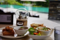 A table set with a variety of dishes including a bowl of soup garnished with herbs, a bowl of ice cream, churros, and fresh vegetables. A small glass of sauce is also present. In the background, there is a blurry view of an outdoor area with greenery and a pool, giving a relaxing ambiance.