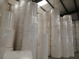 Large rolls of paper are stacked in a warehouse setting with high ceilings and exposed beams. The rolls are uniformly white and cylindrical, arranged in tall columns.