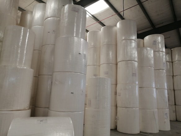 Large rolls of paper are stacked in a warehouse setting with high ceilings and exposed beams. The rolls are uniformly white and cylindrical, arranged in tall columns.