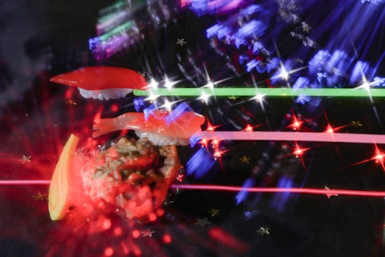 A vibrant and artistic arrangement featuring sushi pieces, such as nigiri and a sushi roll, set against a dark background with colorful light effects. The light creates a dynamic and abstract appearance, with streaks of red, blue, and green and sparkling star-like points.